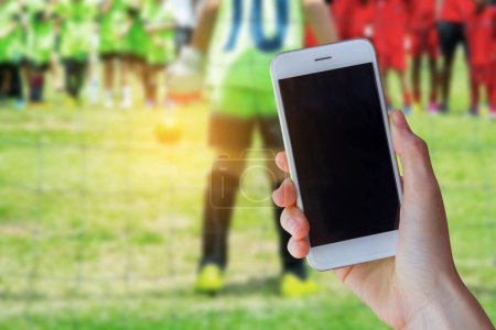 Photo for Hand holding smartphone with young boy as a soccer goalkeeper during football match ready to save. - Royalty Free Image