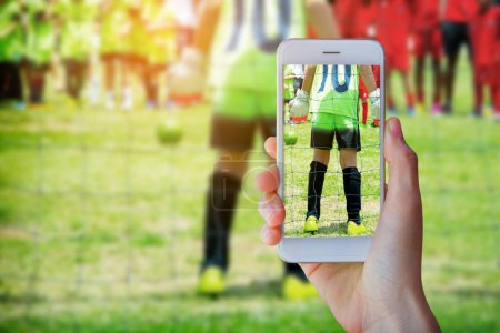 Photo for Hand holding smartphone with young boy as a soccer goalkeeper during football match ready to save. - Royalty Free Image