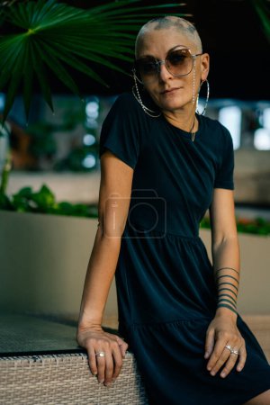 Una joven seria sin pelo en un vestido negro y gafas de sol, mira hacia otro lado. Foto de alta calidad