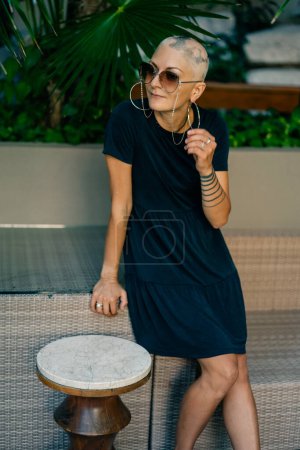 A serious young girl with no hair in a black dress and sunglasses, looks away. High quality photo