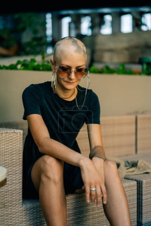 A serious young girl with no hair in a black dress and sunglasses, looks away. High quality photo