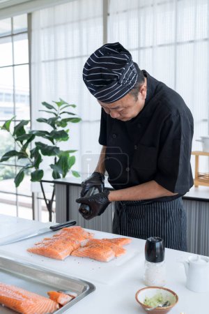Asiatischer Koch mit Salz und schwarzem Pfeffer auf Lachs. Mann schneidet rohen Lachs und bestreut ihn mit schwarzem Pfeffer.