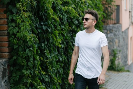 Téléchargez les photos : Hipster beau modèle masculin portant un t-shirt blanc vierge avec de l'espace pour votre logo ou design dans un style urbain décontracté - en image libre de droit