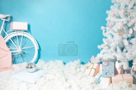 Photo for Christmas shooting location with white christmas tree decorated with toys, present boxes, snow and bike. Artificial fir tree on blue background with copy space, place for text. New year, holiday. - Royalty Free Image