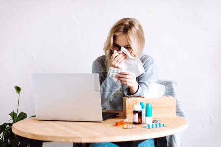 Photo for Portrait of young woman sitting near laptop, different bottles, wooden box with paper napkins, showing pills blister to screen, wiping running nose with napkin, consulting doctor online. Health care. - Royalty Free Image