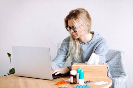 Photo for Portrait of young woman sitting near laptop, bottles with blue caps, box with paper napkins, pills blister, consulting, typing, taking prescription. Telemedicine, online healthcare, online doctor. - Royalty Free Image