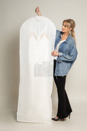 Photo for Vertical blond woman in denim jacket holding and presenting white wedding dress in protective case. Wedding, bride salon and store. Sales for customers, advertisement and marketing. Copy space, mockup - Royalty Free Image
