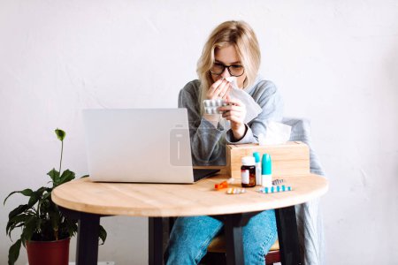 Photo for Portrait of young woman sitting near laptop, bottles with blue caps, wooden box with paper napkins, showing pills blister to screen, wiping running nose, sneezing having video call with doctor online. - Royalty Free Image