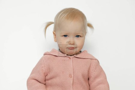 Foto de Nice contented baby girl isolated on white. Portrait of amused toddler in studio. Blonde haired child with pleased facial expression in pink knitted sweater. Beautiful kids concept. Childhood. - Imagen libre de derechos