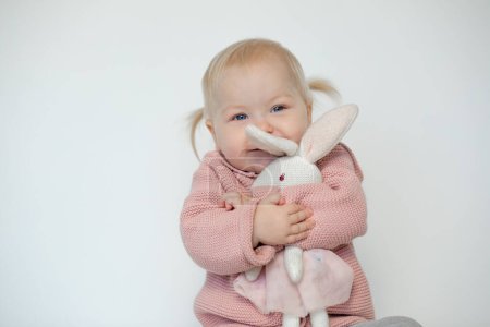 Foto de Beautiful baby girl play with stuffed animal, isolated on white. Joyous toddler happily embracing teddy bunny. Blonde haired child in pink clothes tightly hold paschal rabbit. Easter. Holiday gift. - Imagen libre de derechos