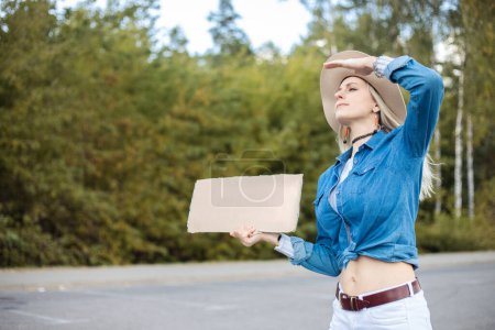Photo for Portrait of young confident blond woman wearing blue shirt, white jeans, beige floppy hat, holding blank cardboard, raising hand to cover eyes from sun, looking narrowly. Travelling, hitchhiking. - Royalty Free Image