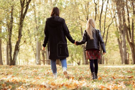 Photo for Back view of family of young woman mother and teenage girl daughter walking on yellow fallen leaves among trees in forest park in autumn, holding hands, relaxing. Relationship, childhood, motherhood. - Royalty Free Image