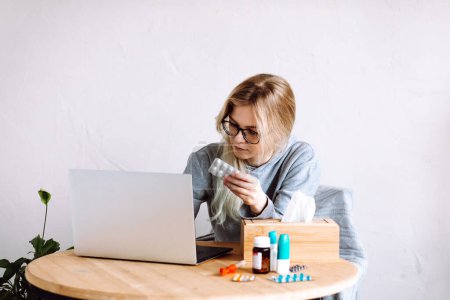 Photo for Portrait of young woman sitting near laptop, bottles with blue caps, box with paper napkins, showing pills blister to screen, consulting, listening to doctor online, taking prescription. Telehealth. - Royalty Free Image