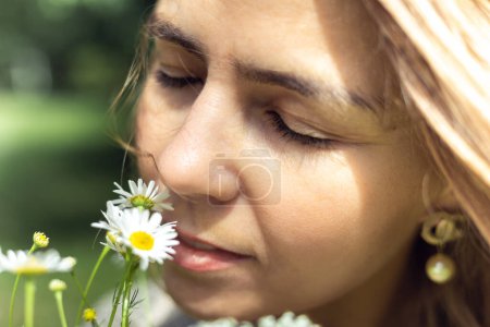 Foto de Close up photo of blond pensive, calm woman holding bouquet of field flowers and smelling it. Unity and harmony with nature. Standing in spring or summer garden, park. Allergy reaction to pollen - Imagen libre de derechos