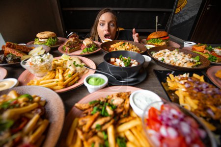 Photo for Hungry woman stare on table full of delicious food in plates with open mouth. Fries, soup, salad, burgers, sauces on festive dinner. Food, many dishes, menu of restaurant, beautiful serving food. - Royalty Free Image