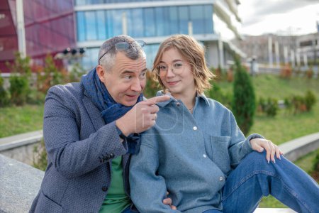 Photo for Smiling mature man explaining and showing something to young woman, sit on railing against university building. Father and daughter, university entrance, enrollee, university entrant, parent support - Royalty Free Image