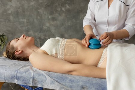 Photo for Cropped photo of masseuse doctor woman applying blue silicone vacuum cups on bare belly of young woman wearing beige underwear, lying on couch in spa salon. Cupping therapy, anti-cellulite massage. - Royalty Free Image