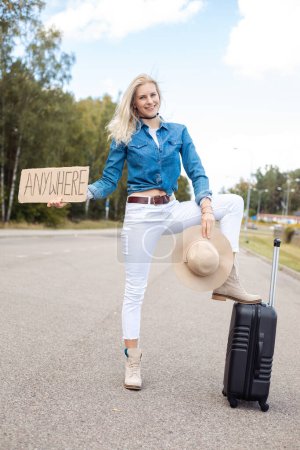 Photo for Vertical positive, laughing, calm blond woman autostopping, show cardboard plate anywhere with raised leg on luggage suitcase and wait for car on highway road. Take me anywhere, travelling abroad - Royalty Free Image