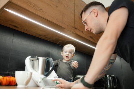 Téléchargez les photos : Man touch settings panel to turn on electronic blender. Little child help dad cooking dinner. Dissatisfied baby son sitting on kitchen tabletop near modern appliances. Family cooking, recipe. - en image libre de droit