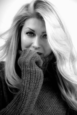 Téléchargez les photos : Attractive 55 years old sexual woman seductively touching face. Female black and white portrait closeup. Beautiful woman with loose thick blonde hair in warm wool sweater. Stylish fashion clothing. - en image libre de droit
