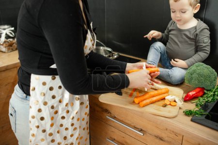 Foto de Mother with little child together cook dinner. Woman peel fresh raw carrots on wooden kitchen board. Ripe organic ingredients, vegetables, healthy nutrition. Family cooking, lifestyle. - Imagen libre de derechos