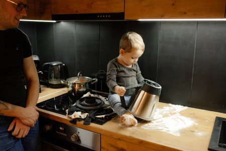 Photo for Smiling man in glasses with crossed hands looking at little blond toddler son with flour on face and clothes playing and holding steel mixer jar, sit on wooden drawer. Adopted child, foster father - Royalty Free Image