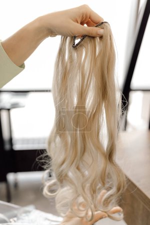 Cropped photo of woman hairdresser hairstylist beautician holding long fair blond hairpiece wig chignon weave bundle for extension in beauty salon. Haircare, hair coloring, dye, hairstyling. Vertical.
