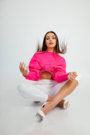 Photo for Stylish woman in pink crop pullover, pants and high heel shoes sitting on floor with flying hair. Full length studio photo, white background. Young woman in sport casual wear. Fashion, basic clothes. - Royalty Free Image