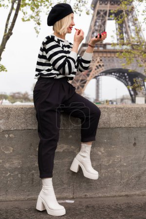 Photo for Adult blonde in black beret, striped blazer sitting on stone fence and painting lips with red lipstick on background of Eiffel Tower. Lady with white high heels shoes and short hair primping outdoor. - Royalty Free Image