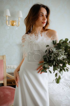 Charming young bride dressed in shoulderless wedding gown adorned with small feathers standing in sleeping room near bed. Dark-haired lady holding bouquet of blossoms in hand look away.