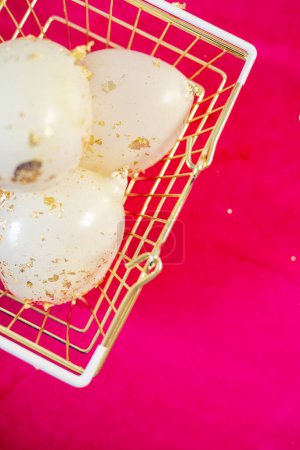 Téléchargez les photos : Vue du dessus du panier à main doré avec des ballons blancs confettis dorés à l'intérieur reposant sur un sol rose vif. Trois bulles élastiques ovales en pannier sur fond rose. Vertical. - en image libre de droit