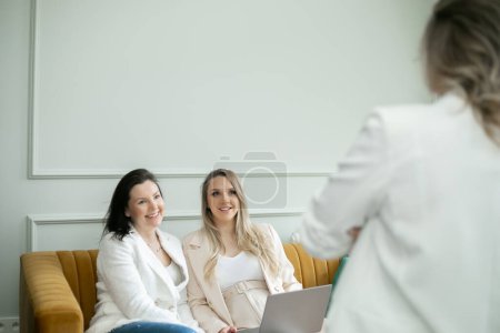Photo for Adorable female team discussing work details in home office. Brunette and preggers lady with laptop sitting on yellow sofa and fervently listening to colleague that standing in front of women. - Royalty Free Image