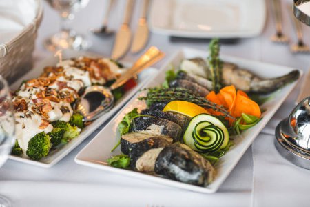 Rechteckige weiße Porzellanteller mit kalten Fischsnacks und Gemüse stehen auf einem schön gedeckten Tisch im Restaurant, Café. Genießen Geschmack. Menü für Feiertage, Hochzeiten, Partys. Kalte Snacks.