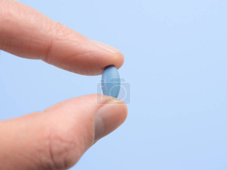 Téléchargez les photos : Pilule bleue pour la dysfonction érectile tenue entre le doigt et le pouce avec un fond bleu. - en image libre de droit