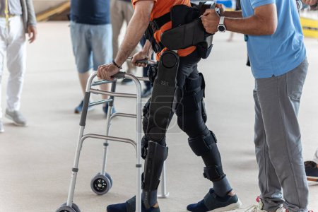 Photo for Boy with Mobility Problems in Lower Limbs in Upright Posture Walking Thanks to a Mechanical Exoskeleton and with the Help of his Physical Therapist. - Royalty Free Image
