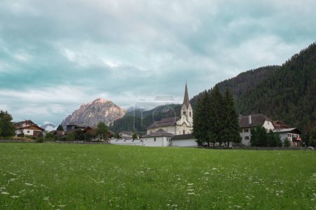 Prados verdes, casas de madera e iglesia en el pueblo de San Vigilio di Marebbe y las montañas italianas de los Alpes en el fondo en un día nublado del verano, Italia.