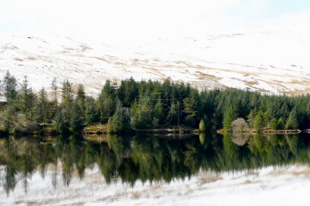 Winter landscape and reflections in the water of the Welsh reservoir of Llwyn Onn, in the Brecon Beacons National Park show the icy mountain side covered in snow and ice and the cloudy sky above. 