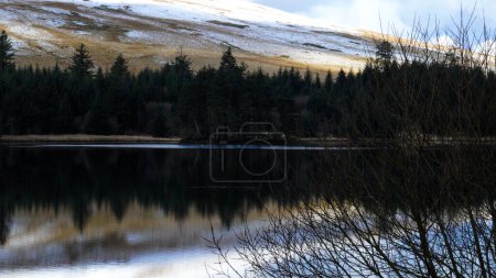 Winter landscape and reflections in the water of the Welsh reservoir of Llwyn Onn, in the Brecon Beacons National Park show the icy mountain side covered in snow and ice and the cloudy sky above. 