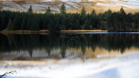 Photo for Winter landscape and reflections in the water of the Welsh reservoir of Llwyn Onn, in the Brecon Beacons National Park show the icy mountain side covered in snow and ice and the cloudy sky above. - Royalty Free Image