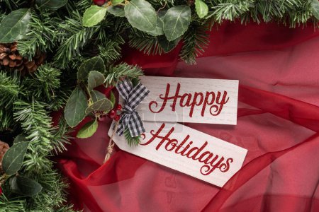Happy Holidays  text on wooden board with sheer red fabric, greenery, and a small black and white gingham bow