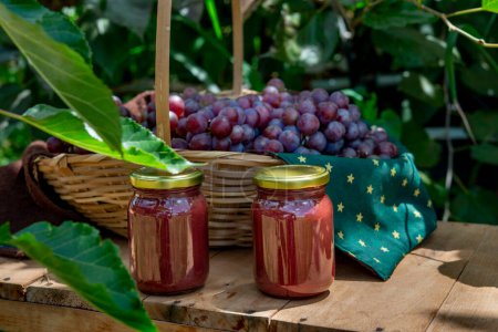 Photo for Grape jam in glass jars with basket full of fresh grapes in background on a wooden table in the garden, jars without label - Royalty Free Image