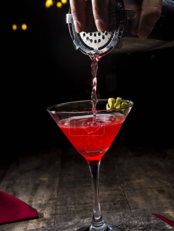 Foto de Cóctel rojo en una copa de martini sobre una mesa de madera con una servilleta roja sobre un fondo negro con un camarero - Imagen libre de derechos