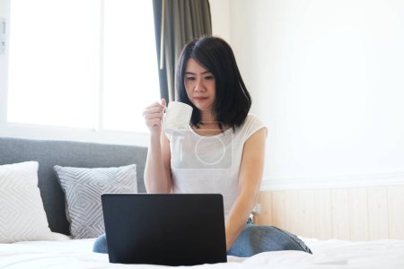 Asiatische Geschäftsfrau trinkt Kaffee und arbeitet mit Laptop im Schlafzimmer. Menschen kaufen mit Technologie online in sozialen Medien ein