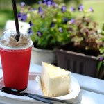 Fresh iced coffee in red glass with cheese cake and cream on wooden bench in flower garden of cafe and coffee shop