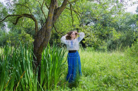Photo for A girl in a long blue skirt and an embroidered shirt among the green meadow and marsh grass and willow trees. - Royalty Free Image