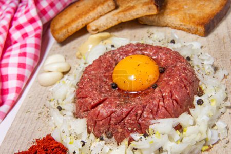 Close-up of tasty raw beef steak tartare with garlic, egg yolk, toasted bread, mustard, ketchup, peppers and onions - a typical variant of Czech cuisine. Checkered red kitchen towel.