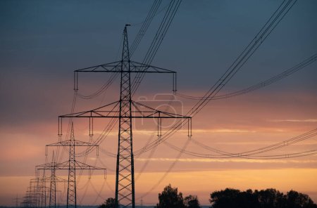 Close-up of large overhead power lines carrying electricity over long distances. The sun rises and shines in the background.