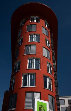Photo for A tall brown-red house in the shape of a tower stands in Augsburg. The building is a dormitory with many small apartments. In the foreground is the advertising sign of an organic food store. - Royalty Free Image