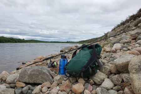 Photo for Kiiruna, Sweden - July 18, 2022: Backback, water bottle with filter and fishing tackle by Lainio river in Swedish Lapland on overcast summer day. - Royalty Free Image