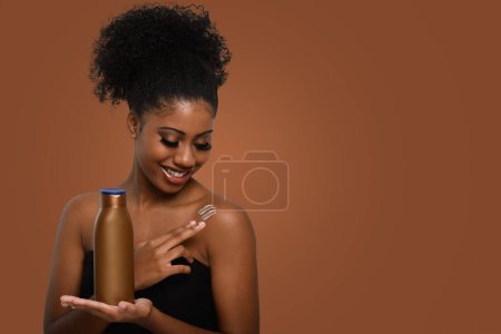 a woman applying moisturizing cream to her skin, smiling while holding a bottle demonstrating skin care, isolated in a brown background